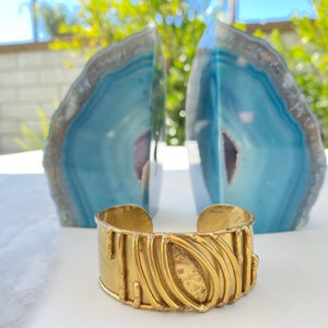 May include: A gold-toned metal cuff bracelet with a textured design. The bracelet has a wide band with a raised oval shape in the center. The bracelet is sitting on a white surface in front of two blue agate bookends.