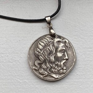 Könnte beinhalten: Silberne Halskette mit Anhänger, der ein detailliertes Profil eines Mannes mit Bart und Krone zeigt. Der Anhänger ist an einer schwarzen Schnur befestigt.