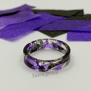 May include: A clear resin ring with purple and black accents. The ring is circular and features embedded pieces of purple and black material. The background includes purple and black paper rectangles.