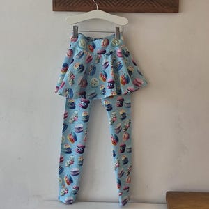 May include: Pale blue leggings and a matching skirt, decorated with a pattern of colourful macarons. The garment hangs on a white hanger. A wooden shelf with a plant and hooks is above.