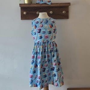 May include: A light blue dress featuring a pattern of colourful macarons, with a matching headband. The dress is displayed on a mannequin. A wooden shelf with brass knobs and a potted plant are in the background. The dress is approximately 90 cm in length.