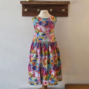 Könnte beinhalten: Ein ärmelloses, farbenfrohes Kleid mit einem lebendigen Muster aus Regenbogen, Blumen und Herzen. Das Kleid wird auf einer Schaufensterpuppe präsentiert. Im Hintergrund befinden sich ein Holzregal mit Messinghaken und eine Topfpflanze.