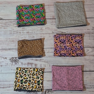 May include: Six neck gaiters with different animal print designs are arranged on a wooden surface. The patterns include a rainbow leopard print, a grey and white spotted print, a gold glitter print, a purple and orange leopard print, a tan and black leopard print, and a pink spotted print.