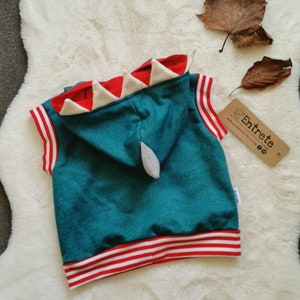 Puede incluir: Sudadera azul turquesa sin mangas con ribete a rayas rojas y blancas. La sudadera tiene una capucha en forma de dinosaurio roja y blanca con dientes blancos.