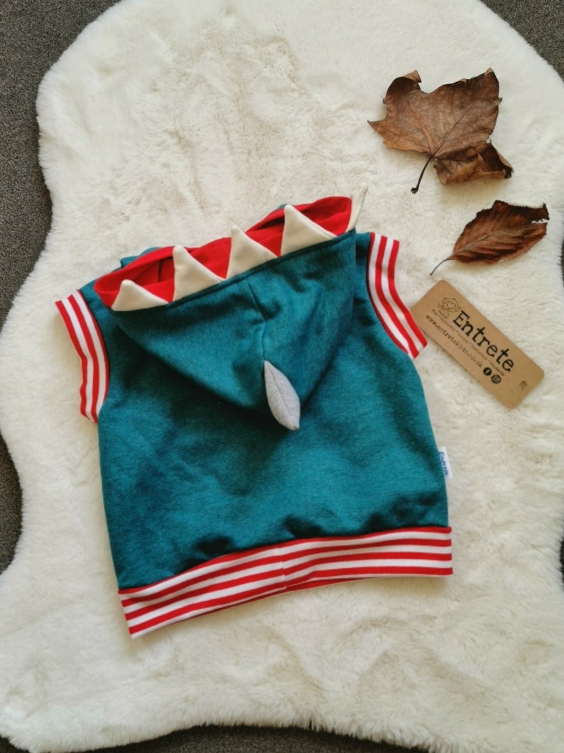 Puede incluir: Sudadera azul turquesa sin mangas con ribete a rayas rojas y blancas. La sudadera tiene una capucha en forma de dinosaurio roja y blanca con dientes blancos.