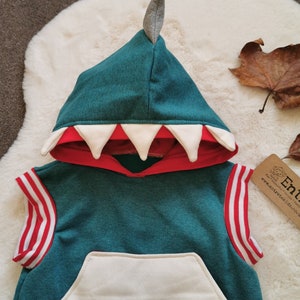 Puede incluir: Una sudadera con capucha de color turquesa sin mangas con un bolsillo blanco y rayas rojas y blancas en las mangas. La capucha tiene un dise&ntilde;o de dientes de dinosaurio blanco y rojo y una punta de fieltro gris en la parte superior.