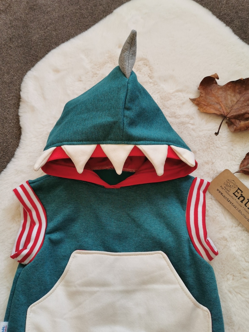 Puede incluir: Una sudadera con capucha de color turquesa sin mangas con un bolsillo blanco y rayas rojas y blancas en las mangas. La capucha tiene un dise&ntilde;o de dientes de dinosaurio blanco y rojo y una punta de fieltro gris en la parte superior.
