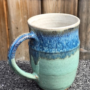 May include: A ceramic mug featuring a two-tone glaze with a deep blue upper section that transitions into a light mint green lower body. The mug has a curved handle with a textured, speckled finish and a smooth, off-white interior surface.