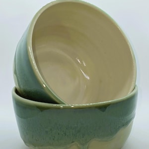 May include: Two ceramic bowls with a green glaze. The bowls are nested together, with the top bowl showing a light yellow interior. The bottom bowl has a light yellow rim.