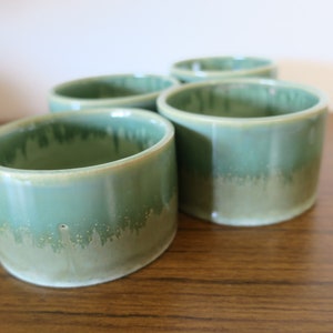 May include: Four green ceramic cups with a light green glaze and a subtle speckled pattern. The cups are arranged in a row on a wooden surface.