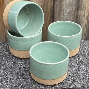 May include: A set of three ceramic cups featuring a smooth, light mint green glazed interior and upper exterior, contrasted by a textured, unglazed tan base. The cups are cylindrical in shape with a wide opening and a speckled finish on the glaze.