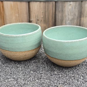 May include: Two ceramic bowls featuring a smooth, light mint green glazed upper section and a textured, unglazed tan clay base. The bowls have a rounded, wide-mouthed shape with a simple, clean design that highlights the contrast between the matte finish of the base and the glossy, speckled appearance of the mint green glaze.