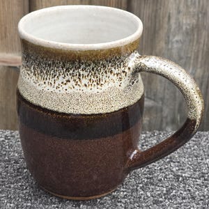 May include: A ceramic mug featuring a two-tone glaze design. The upper portion displays a speckled cream and brown pattern, while the lower section is a solid, deep dark brown. The mug has a curved handle and a smooth, rounded rim.