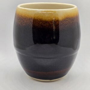 May include: A ceramic mug with a brown, black and cream glaze. The mug has a rounded shape and a slightly flared lip.