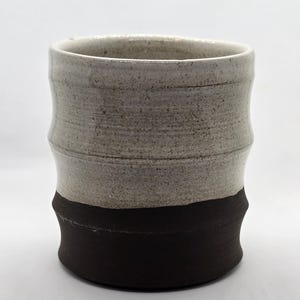 May include: A cylindrical ceramic cup featuring a two-tone design. The upper portion is a speckled light beige, while the lower portion is a solid dark brown. The exterior surface has a subtle, undulating texture with horizontal ridges.
