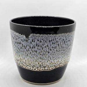May include: A black ceramic planter with a white and brown speckled glaze. The planter has a wide, round opening and a slightly flared lip.