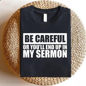 May include: Black t-shirt with the text "BE CAREFUL OR YOU'LL END UP IN MY SERMON" in a white, bold font. The shirt is folded and placed on a woven, circular surface. The t-shirt has a crew neck.
