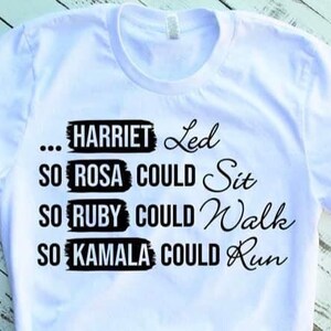 May include: White t-shirt with black text. The text reads: "...HARRIET Led, SO ROSA COULD Sit, SO RUBY COULD Walk, SO KAMALA COULD Run." The shirt is on a light wood grain surface.