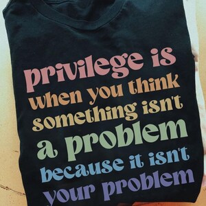 以下が含まれることがあります： 虹色の文字で「privilege is when you think something isn't a problem because it isn't your problem」と書かれた黒いTシャツです。
