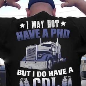 Op de afbeelding: Een zwart T-shirt met een witte afbeelding van een vrachtwagen en de tekst "I may not have a PHD but I do have a CDL".