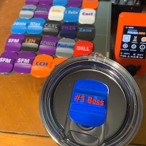 Puede incluir: Primer plano de una tapa impresa en 3D azul y roja con el texto "#1 Boss" en ella. La tapa está encima de un vaso negro y plateado.