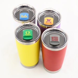 Puede incluir: Cuatro termos de viaje de acero inoxidable con tapas y logotipos de diferentes colores. Las tapas tienen insertos de plástico extraíbles y coloridos con logotipos de equipo y años. Los logotipos son "UCLA", "W 2014", "O 1999" y "WSU 2020".