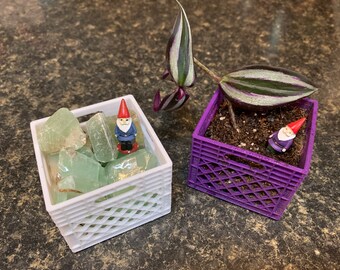 May include: Two small white and purple crates with a gnome figurine in each. The white crate contains green gemstones and the purple crate contains a small green plant with striped leaves.