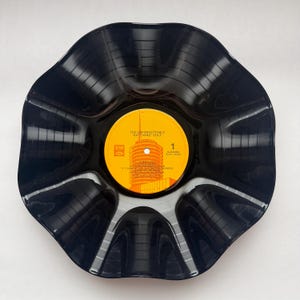 May include: A black vinyl record bowl with a scalloped, wavy edge. The center features a bright orange label with the text The Unforgettable Nat King Cole, SM logo, and a tracklist including Mona Lisa and Nature Boy, with a central hole.