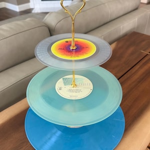 May include: A three-tiered serving stand constructed from stacked vinyl records in clear, light blue, and solid blue colors. The stand features a central gold-toned metal rod with a decorative handle at the top, holding the records in place.