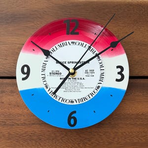 Puede incluir: Un reloj de pared redondo hecho de un disco de vinilo con un fondo de rayas horizontales rojas, blancas y azules. La etiqueta central muestra el texto Bruce Springsteen Born in the U.S.A. con manecillas negras y números grandes.