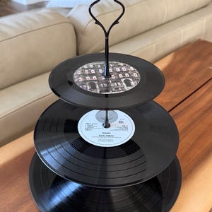 May include: A three-tiered serving stand constructed from black vinyl records. The top tier features a Physical Graffiti label, while the middle tier displays a Black Sabbath Paranoid Vertigo label. A black metal handle connects the tiers.