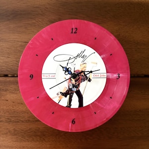 May include: A round wall clock made from a pink marbled vinyl record. The center features an image of a person holding tools with the text Dolly, 9 to 5 and Odd Jobs. The clock has black hour, minute, and second hands with black numerals at 3, 6, 9, and 12.