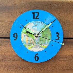 May include: A round wall clock crafted from a blue vinyl record featuring the Fleetwood Mac Rumours album label in the center. The clock has large black numerals at the 3, 6, 9, and 12 positions, with thin black hands tipped in light green.