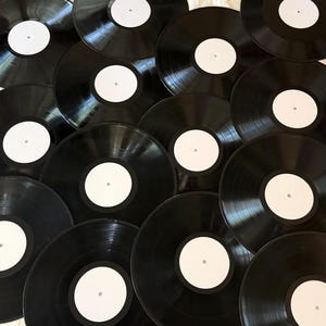 May include: A collection of multiple black vinyl records arranged in an overlapping pattern. Each circular record features a smooth, glossy black surface with a plain, solid white circular label in the center, showing no text or graphics.