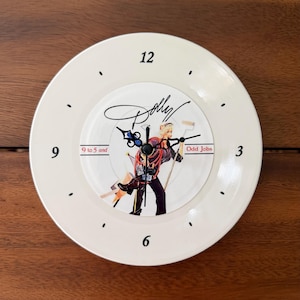 May include: A round wall clock with a cream-colored face featuring black numerals at 3, 6, 9, and 12. The center displays an illustration of a person with the text Dolly, 9 to 5 and Odd Jobs, and black ornate clock hands.