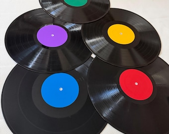 May include: Five black vinyl records arranged in a cluster. Each record features a solid-colored center label in green, purple, yellow, blue, or red. The circular discs have visible grooves and a central hole, showcasing a glossy finish on the black surface.