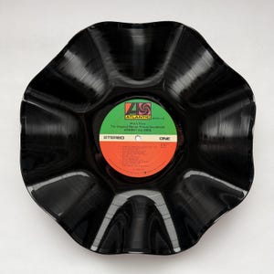 May include: A black, upcycled vinyl record bowl. The center label is green and orange, with the text "ATLANTIC" and "AGAINST ALL ODDS". The bowl has a wavy, petal-like shape, and is a unique decorative item.