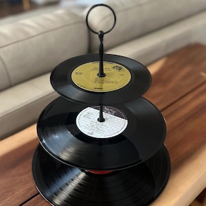 以下が含まれることがあります： 黒いビニールレコードで作られた3段のサービングスタンド。黒いレコードは、黒い金属製のロッドと上部の円形のハンドルで積み重ねられています。ラベルは黄色と白です。