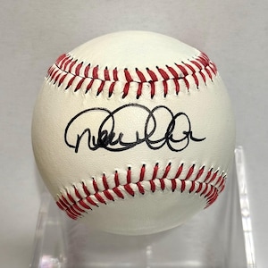 Puede incluir: Una pelota de béisbol blanca con costuras rojas y un autógrafo en tinta negra en el centro. La pelota es esférica con una superficie de cuero texturizado y está colocada para mostrar claramente la firma sobre las costuras rojas.