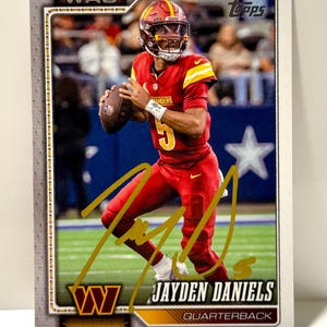 May include: A Topps football trading card featuring Jayden Daniels in a red Washington Commanders uniform, holding a football. The card includes a gold ink autograph across the centre, the text WAS, the player name Jayden Daniels, and Quarterback.