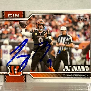 May include: A Topps trading card featuring Joe Burrow, the Cincinnati Bengals quarterback, in a black uniform with the number 9. The card displays a blue ink autograph across the center, the Topps logo, and the text CIN and JOE BURROW QUARTERBACK.