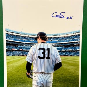 Può includere: Una fotografia lucida che ritrae un giocatore di baseball di spalle, che indossa una maglia bianca a righe con il numero 31 sulla schiena, in piedi su un campo da baseball. L'immagine include un autografo a inchiostro blu e il numero 31 in alto.