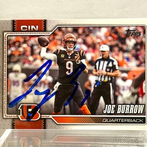 May include: A Topps trading card featuring Joe Burrow in a black Cincinnati Bengals uniform with the number 9, throwing a football. The card includes a blue ink autograph across the front, the text CIN, Joe Burrow, and Quarterback.