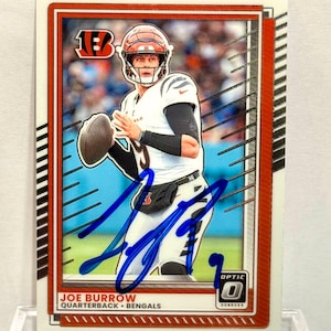 May include: A rectangular sports trading card featuring Joe Burrow in a white Cincinnati Bengals uniform, holding a football. The card has an orange border, a blue ink autograph across the center, and text reading Joe Burrow Quarterback Bengals.