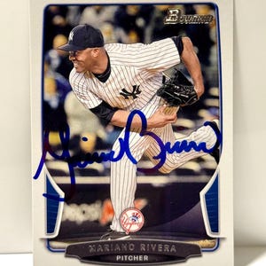 Può includere: Una figurina da baseball rettangolare con una fotografia di Mariano Rivera in divisa bianca gessata dei New York Yankees, durante un lancio. La carta include un autografo a inchiostro blu, il logo Bowman e il testo Mariano Rivera Pitcher.