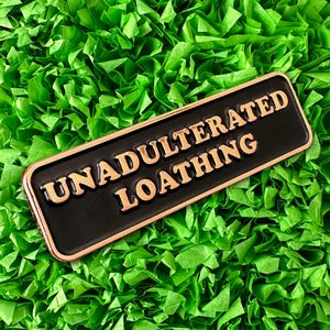 May include: A black and gold enamel pin with the text "UNADULTERATED LOATHING" in gold letters.