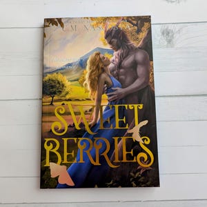 Könnte beinhalten: Ein Taschenbuch mit dem Titel "Sweet Berries" mit einem romantischen Fantasy-Cover. Das Cover zeigt eine muskulöse, geflügelte männliche Figur, die eine Frau in einem blauen Kleid umarmt. Der Titel ist in Gold, mit dem Untertitel "a Monster Butt Romance."