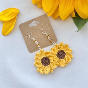 May include: Crocheted sunflower earrings with brown centers and silver hooks. The earrings are displayed on a brown card with a sunflower in the background.