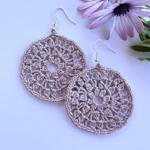 May include: A pair of crocheted earrings in a light brown color. The earrings are round and have a delicate, intricate design. They are hanging from silver hooks.