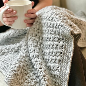 May include: A gray crocheted throw blanket with a textured pattern. The blanket is draped over a person's lap.